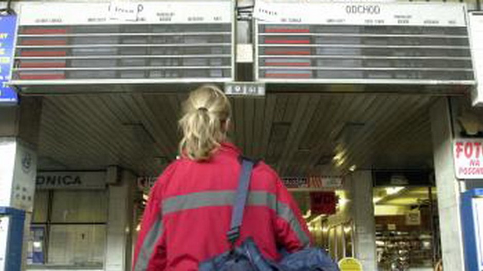 zena na zeleznicnej stanici, za nou tabula s prichodmi vlakov